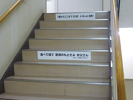 写真：階段の写真には「食べて寝て肥満のもとだよお父さん」などの標語が貼られている。